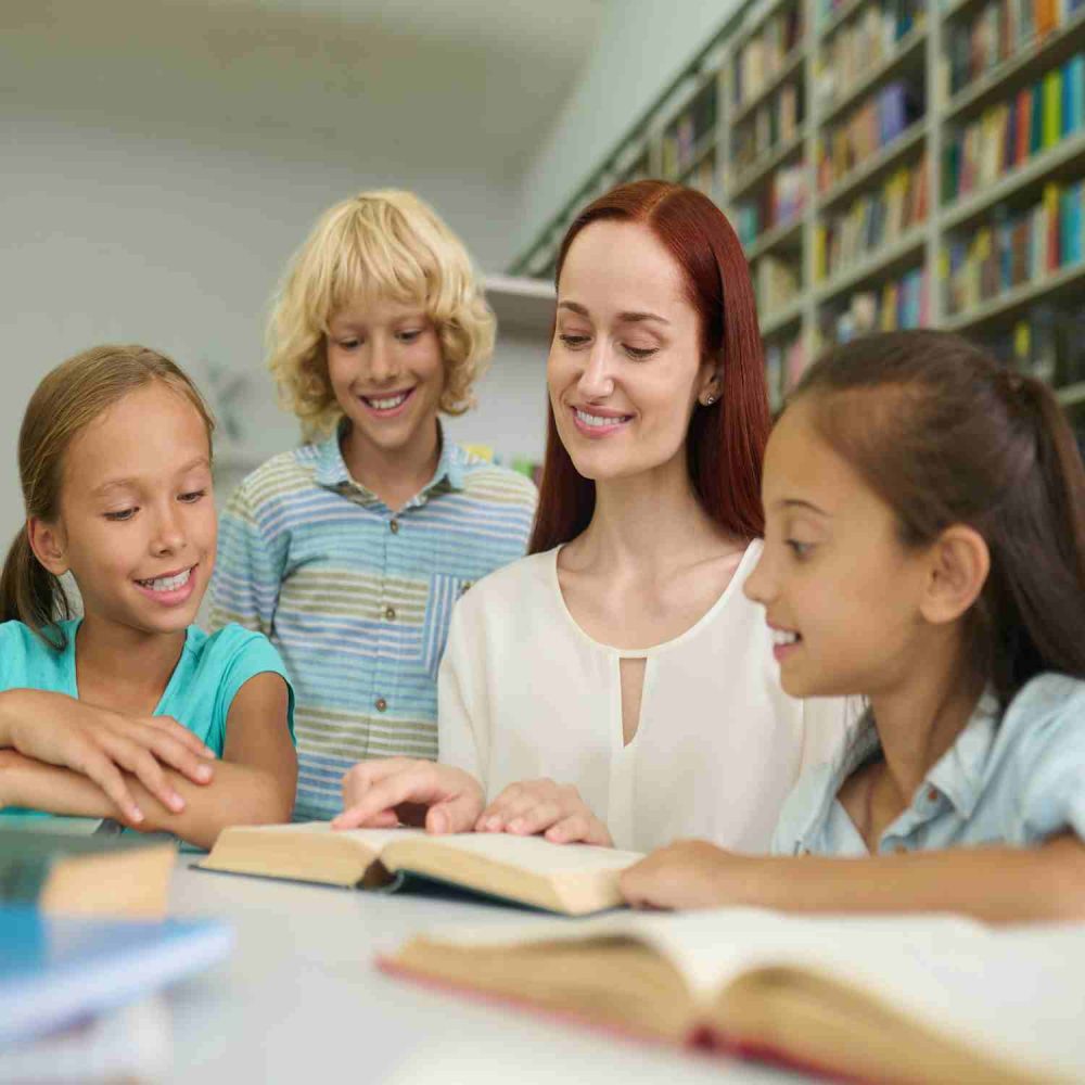 Teamwork. Young smiling woman touching finger to page of open book and happy primary school children reading book together at table in library